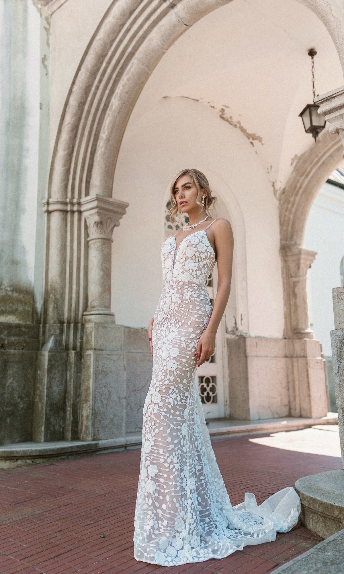 A woman stands outdoors before stone arches, wearing the Long White Lace Formal Dress: Tina Holly BB008, featuring a fitted silhouette with floral lace patterns and sheer design.